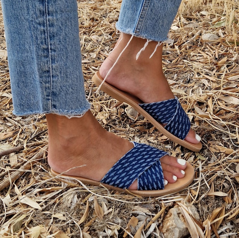 K&ouml;nnte beinhalten: Ein Paar blaue Jeans-Sandalen mit einem gekreuzten Riemen-Design. Die Sandalen haben eine braune Ledersohle und werden an den F&uuml;&szlig;en getragen.