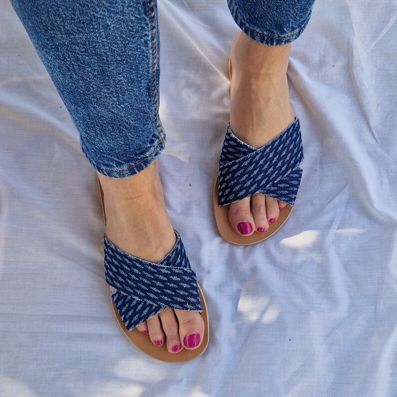 K&ouml;nnte beinhalten: Ein Paar blaue Jeans-Sandalen mit gekreuzten Riemen. Die Sandalen haben eine braune Ledersohle und werden getragen.