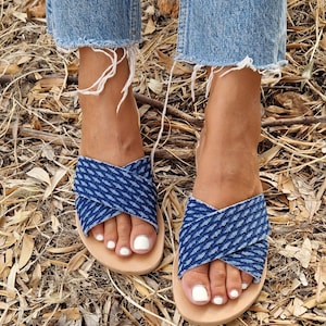 K&ouml;nnte beinhalten: Ein Paar blaue Jeans-Sandalen mit einem gekreuzten Riemen-Design. Die Sandalen haben eine hellbraune Ledersohle und werden getragen.