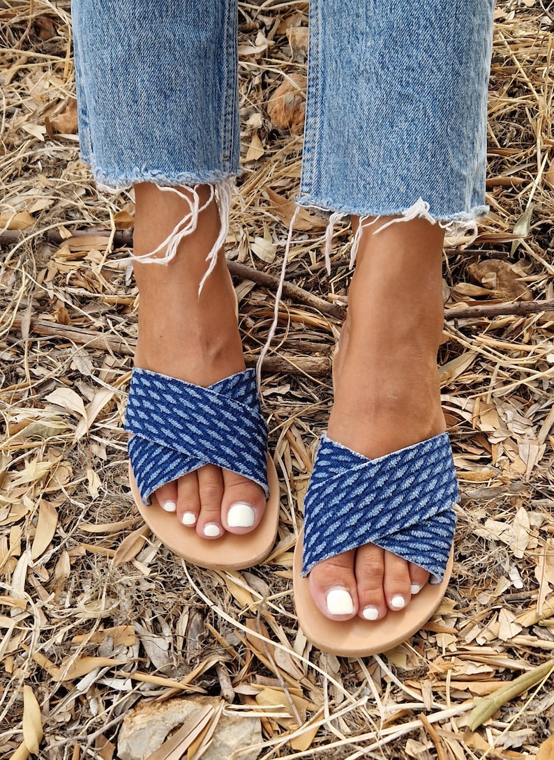 K&ouml;nnte beinhalten: Ein Paar blaue Jeans-Sandalen mit einem gekreuzten Riemen-Design. Die Sandalen haben eine hellbraune Ledersohle und werden getragen.
