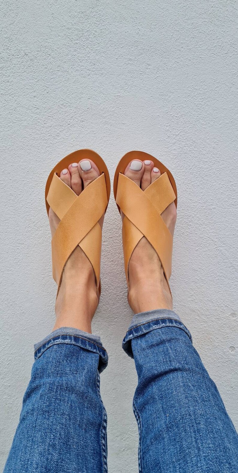 May include: A pair of tan leather sandals with criss-cross straps. The sandals are worn by a person wearing blue jeans.