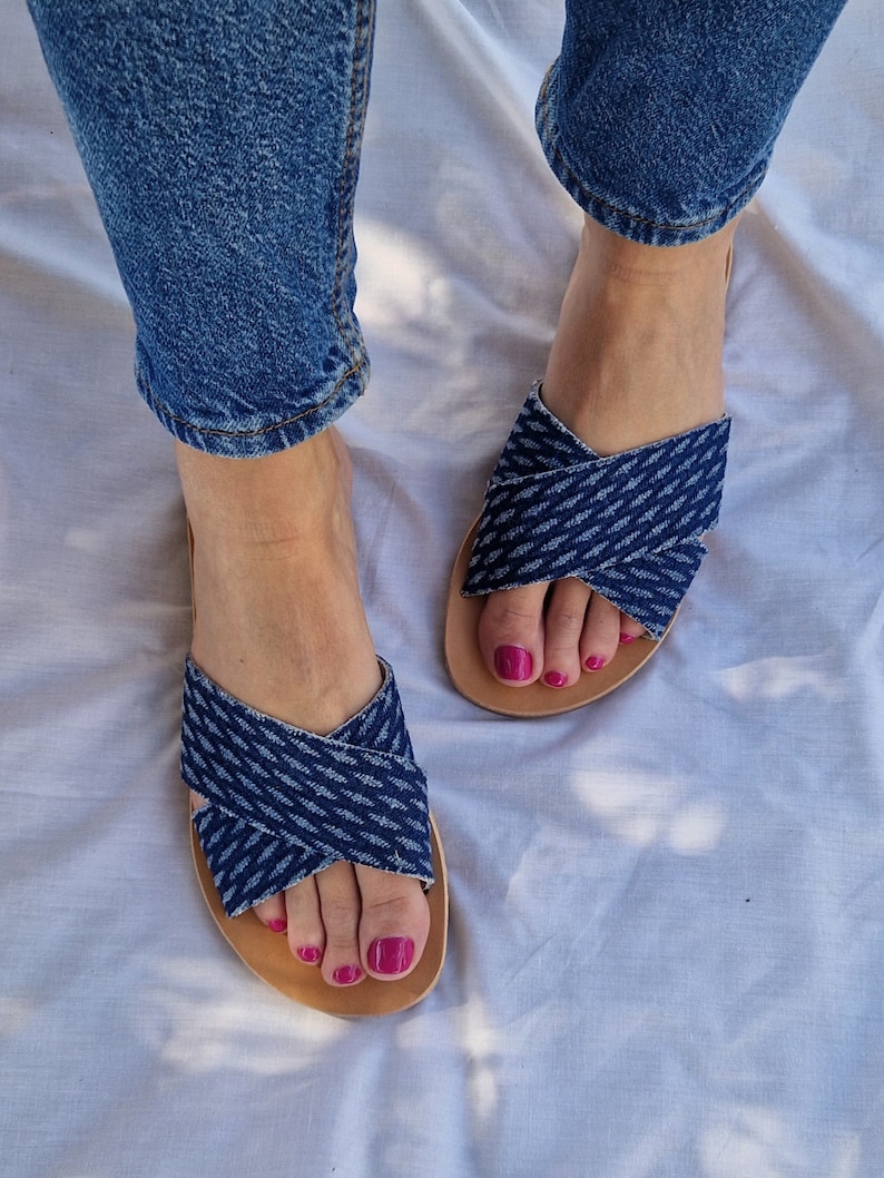 K&ouml;nnte beinhalten: Ein Paar blaue Jeans-Sandalen mit gekreuzten Riemen. Die Sandalen haben eine braune Ledersohle und werden an den F&uuml;&szlig;en einer Person mit rosafarbenem Nagellack getragen.