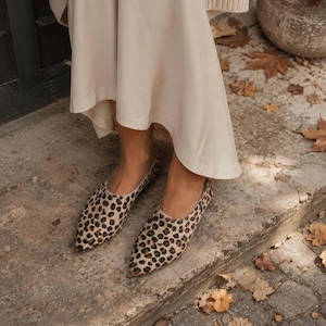 May include: A pair of pointed-toe flats featuring a brown and black leopard print pattern. The shoes have a low-cut topline and a slim silhouette, worn with a long, flowing cream-colored skirt that reaches down toward the ankles.