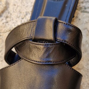 May include: Black leather sandal with a knotted strap design. The sandal has a simple, minimalist style.