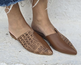 May include: A pair of brown leather mules with pointed toes. One shoe features a textured, crocodile-like pattern on the vamp, while the other has a smooth finish. The shoes are open-backed and sit on a white surface.