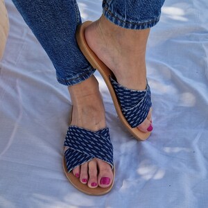 K&ouml;nnte beinhalten: Ein Paar blaue Jeans-Sandalen mit gekreuzten Riemen. Die Sandalen haben eine braune Ledersohle und werden an den F&uuml;&szlig;en einer Person getragen.