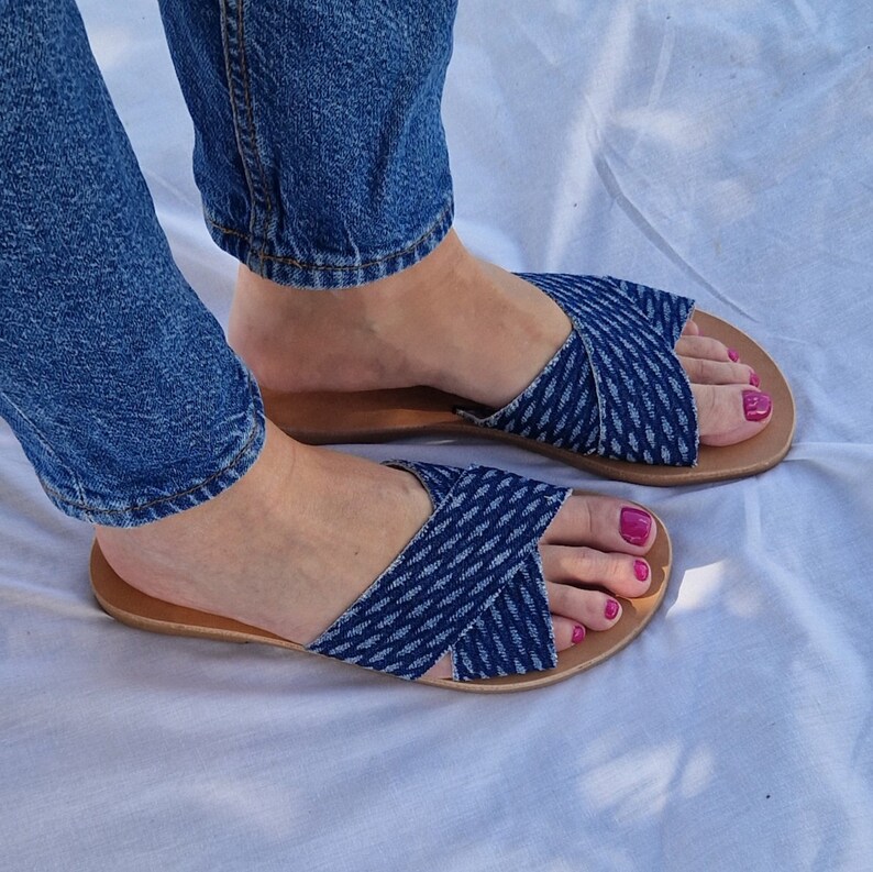 K&ouml;nnte beinhalten: Ein Paar blaue Jeans-Sandalen mit gekreuzten Riemen. Die Sandalen haben eine braune Ledersohle und werden an den F&uuml;&szlig;en einer Person getragen.