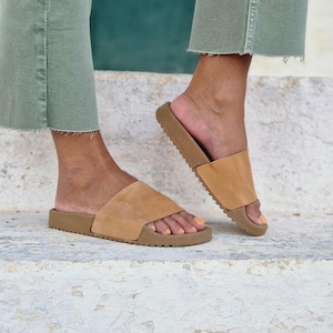 以下が含まれることがあります： 厚いゴム製のソールが付いたタン色の革製のサンダル。サンダルは人の足に履かれている。