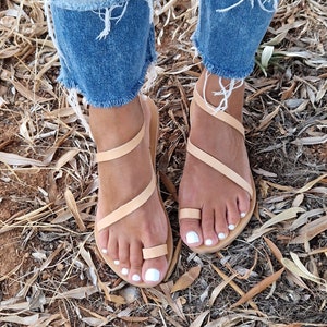 K&ouml;nnte beinhalten: Tannfarbene Ledersandalen mit mehreren Riemen, die sich um die Kn&ouml;chel und Zehen wickeln. Die Sandalen haben ein schlichtes Design und werden barfu&szlig; getragen.