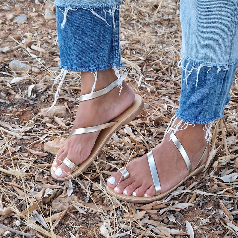 K&ouml;nnte beinhalten: Ein Paar silberne Ledersandalen mit Zehenring und Kn&ouml;chelriemen. Die Sandalen werden von einer Person mit nackten F&uuml;&szlig;en getragen und vor einem Hintergrund aus trockenen, braunen Bl&auml;ttern platziert.