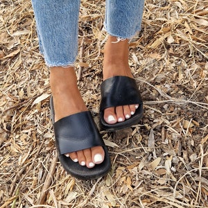 以下が含まれることがあります： シンプルなデザインの黒い革製のサンダル。サンダルはフラットソールで、白いマニキュアを塗った素足で履いています。