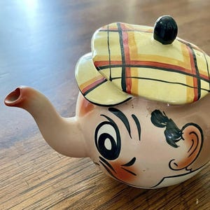 May include: A novelty teapot with a face design, featuring a tan body, a spout, and a handle. The lid is a yellow, red, and black plaid pattern, and the teapot has a black knob. The face has black eyebrows, a mustache, and rosy cheeks.