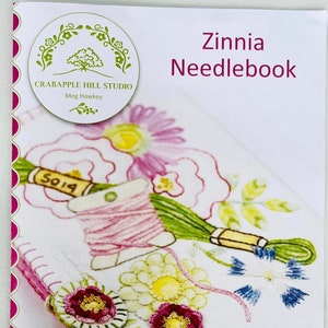 May include: A white needlebook with pink stitching around the edges. The front of the needlebook features an embroidered design of flowers, a spool of thread, and a needle. The text "Zinnia Needlebook" is printed in pink at the top of the needlebook. The text "Crabapple Hill Studio Meg Hawkey" is printed in green at the bottom of the needlebook.
