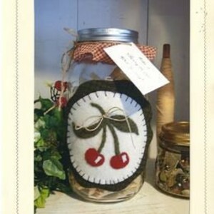 May include: A glass jar with a white felt cherry applique on the front. The jar is decorated with a red and white checkered fabric and a tag that reads "As the Crow Flies..."