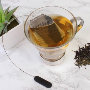 May include: A stainless steel tea infuser with a black silicone handle, hanging in a glass mug filled with tea.