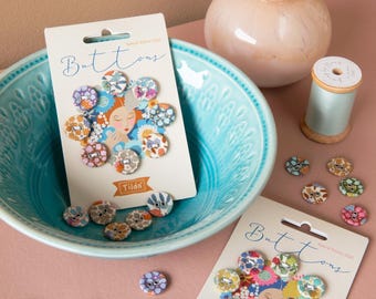 May include: A collection of decorative buttons with floral and animal patterns. The buttons are displayed on cards that read "Buttons" and "Tilda." A light blue bowl and a pink vase with a tulip are also in the image.