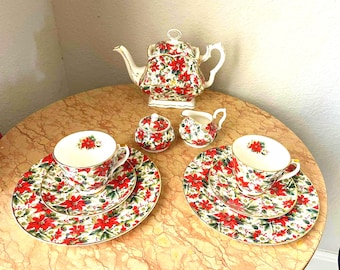 May include: A complete tea set with a floral poinsettia design. The set includes a teapot, sugar bowl, creamer, two cups, and saucers. The cups and saucers are stacked on top of each other. The set is on a round table.