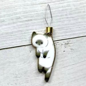 May include: A white cat with black markings hanging from a gold needle threader. The cat is facing to the left and has a surprised expression.