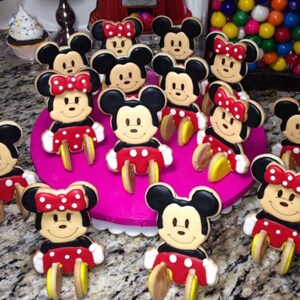 May include: A collection of Mickey and Minnie Mouse-shaped cookies. The cookies feature black ears, smiling faces, red shorts with white polka dots, and yellow shoes. Some cookies have red bows with white polka dots.