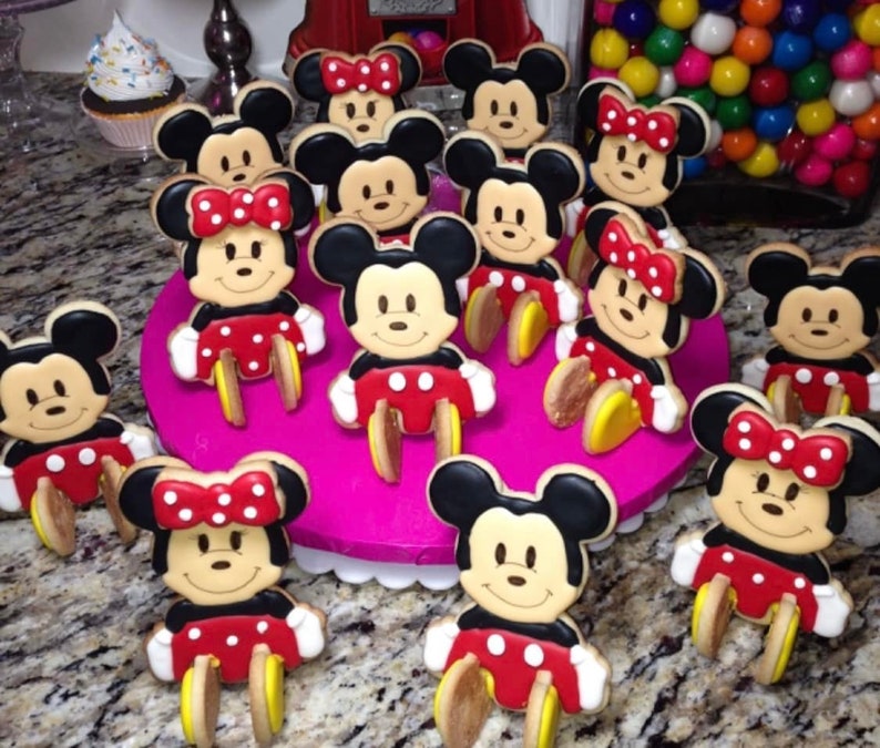 May include: A collection of Mickey and Minnie Mouse-shaped cookies. The cookies feature black ears, smiling faces, red shorts with white polka dots, and yellow shoes. Some cookies have red bows with white polka dots.