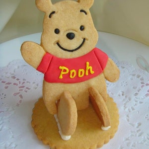 May include: A Winnie the Pooh cookie, featuring a smiling bear with a red shirt and the word "Pooh" in yellow. The cookie is on a round, scalloped base, with the bear's legs extending down. The cookie is light brown.