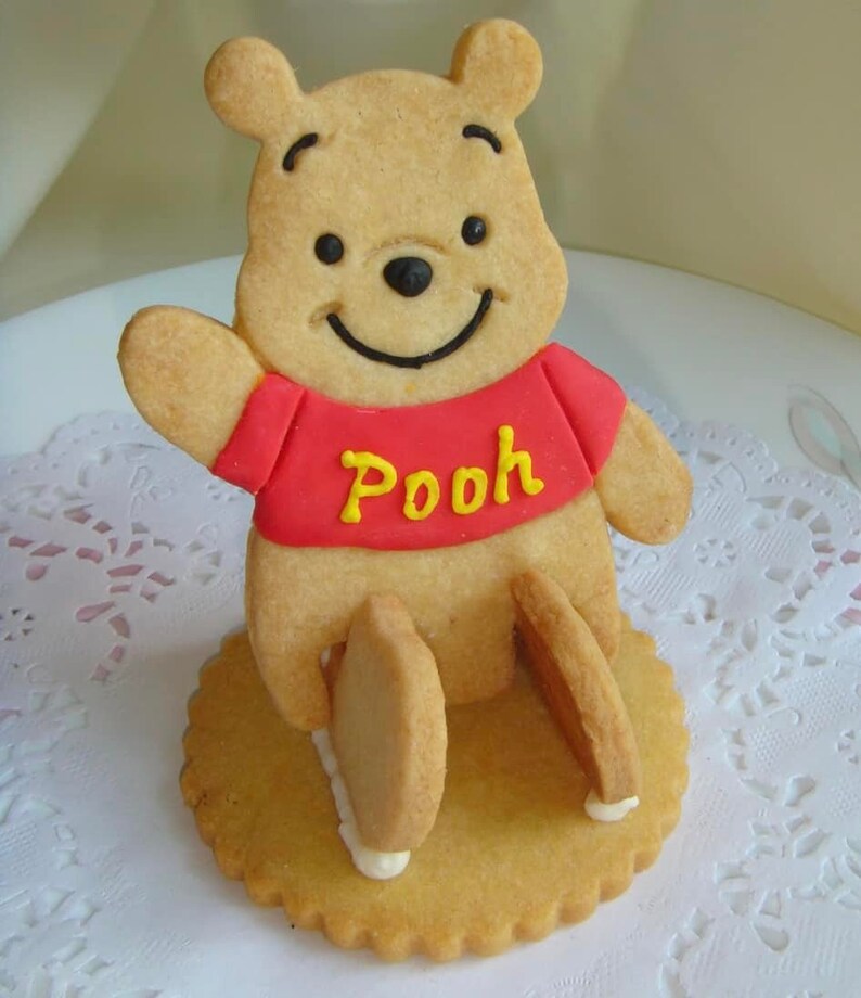 May include: A Winnie the Pooh cookie, featuring a smiling bear with a red shirt and the word "Pooh" in yellow. The cookie is on a round, scalloped base, with the bear's legs extending down. The cookie is light brown.