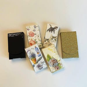May include: An assortment of decorative matchbox holders. One is black and glossy, another is gold glitter, and the others feature floral and cat-themed designs. The matchbox holders are rectangular with a curved top.