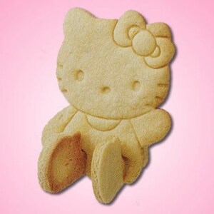 May include: A light-colored, baked cookie in the shape of Hello Kitty. The cookie features a detailed design of the character's face, bow, arms, and legs. The cookie is set against a pink background.