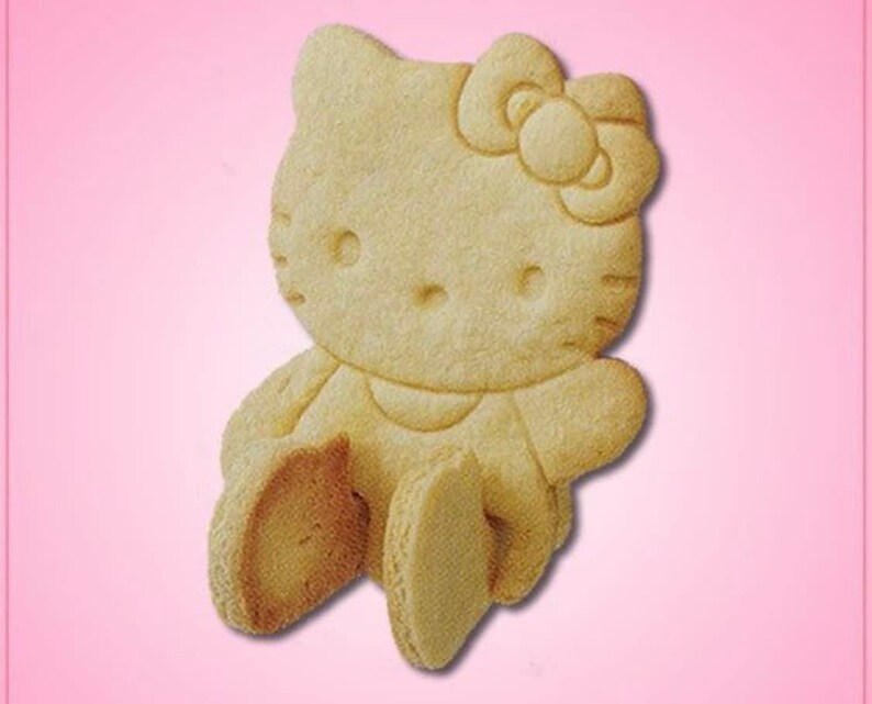 May include: A light-colored, baked cookie in the shape of Hello Kitty. The cookie features a detailed design of the character's face, bow, arms, and legs. The cookie is set against a pink background.