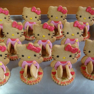 May include: A tray of decorated cookies shaped like the character Hello Kitty. Each cookie has a pink bow, dress, and a base with the words "Happy 1st Birthday". The cookies are arranged on a metal tray.