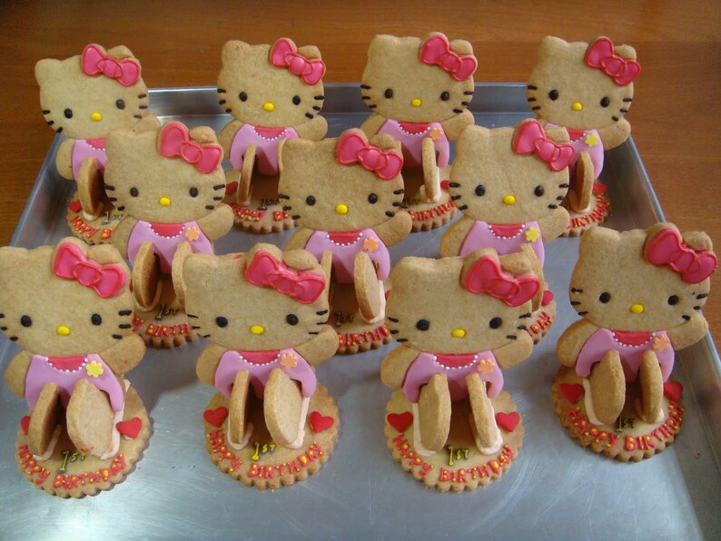 May include: A tray of decorated cookies shaped like the character Hello Kitty. Each cookie has a pink bow, dress, and a base with the words "Happy 1st Birthday". The cookies are arranged on a metal tray.
