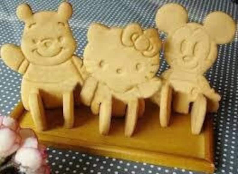 May include: Three cartoon character cookies: Winnie the Pooh, Hello Kitty, and Mickey Mouse, sit on a rectangular wooden tray. The cookies are light brown and have detailed facial features and body outlines. The tray is a darker shade of brown.