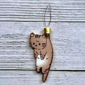 May include: A brown and white cat charm with a gold loop for hanging. The cat is hanging upside down with its tail pointing down.