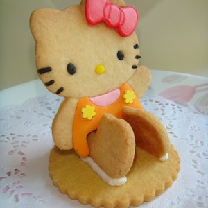 May include: A Hello Kitty-shaped cookie with a pink bow, black eyes, and an orange dress with yellow flower accents. The cookie is on a round, scalloped-edge cookie base, with white icing accents.