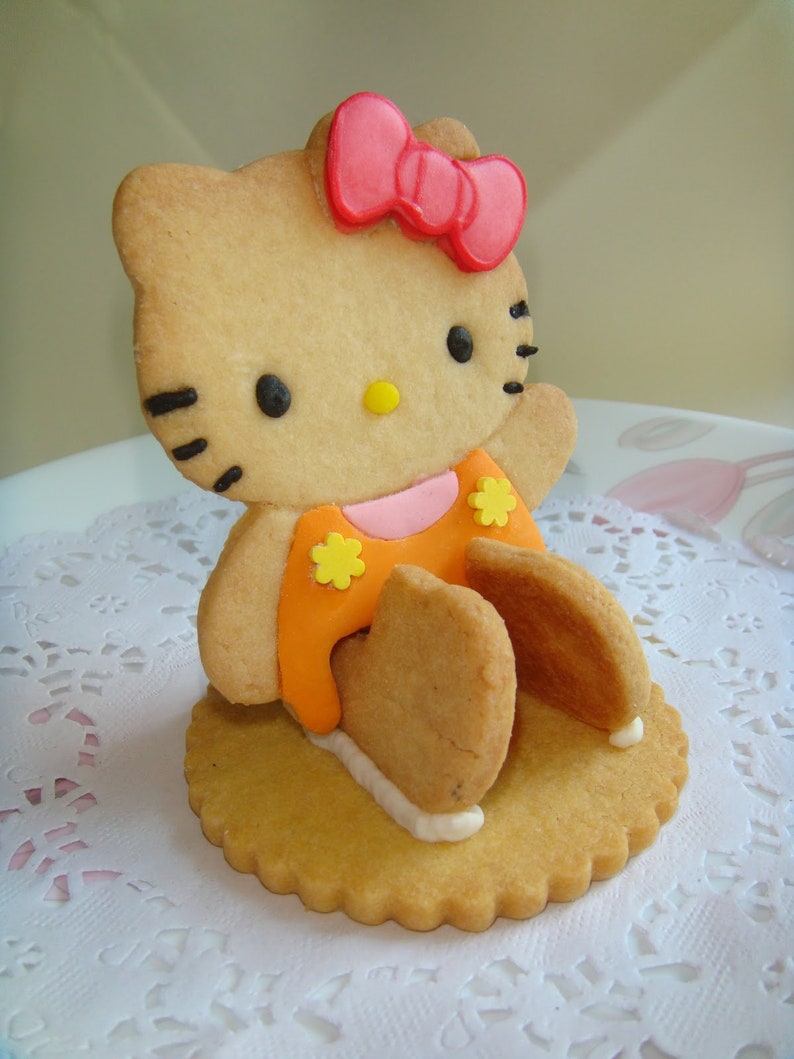 May include: A Hello Kitty-shaped cookie with a pink bow, black eyes, and an orange dress with yellow flower accents. The cookie is on a round, scalloped-edge cookie base, with white icing accents.