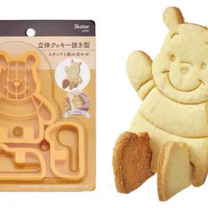 May include: A Winnie the Pooh cookie cutter set and a baked Winnie the Pooh cookie. The cookie cutter is orange plastic and includes the Disney logo. The cookie is light yellow with a smiling face.