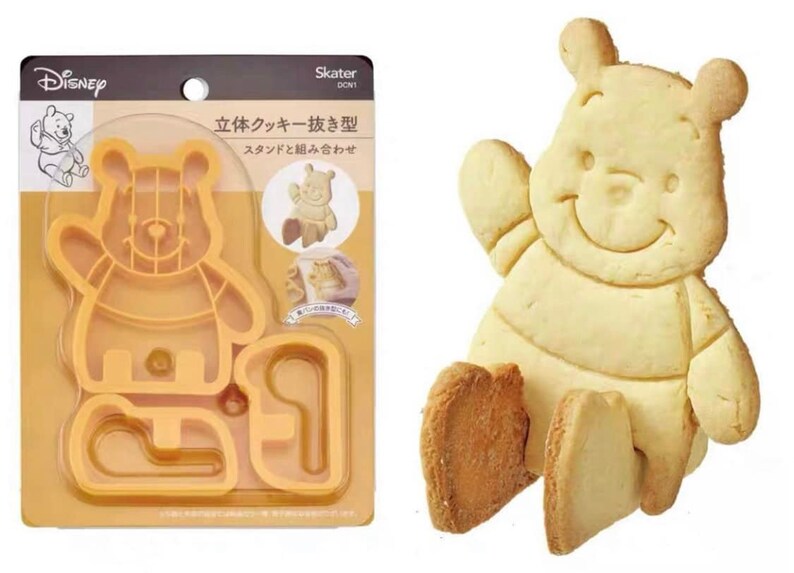 May include: A Winnie the Pooh cookie cutter set and a baked Winnie the Pooh cookie. The cookie cutter is orange plastic and includes the Disney logo. The cookie is light yellow with a smiling face.