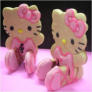 May include: Two Hello Kitty-shaped cookies with pink bows, dresses, and ballet slippers. The cookies have black eyes, whiskers, and yellow noses. One cookie has a musical note on its dress. The cookies are on a pink surface.