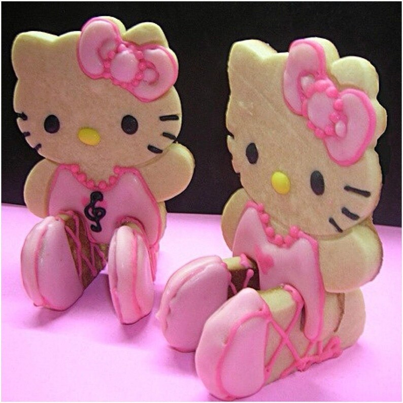 May include: Two Hello Kitty-shaped cookies with pink bows, dresses, and ballet slippers. The cookies have black eyes, whiskers, and yellow noses. One cookie has a musical note on its dress. The cookies are on a pink surface.