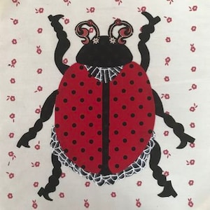 May include: A fabric applique of a ladybug with a red body covered in black dots. The ladybug has black legs, antennae, and a black head. The ladybug is decorated with a white lace trim and is set against a white background with small red floral patterns.