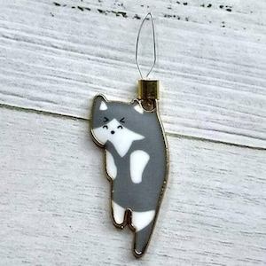 May include: A gray and white cat-shaped needle threader with a gold metal loop. The cat has a white face with black eyes and a black mouth.