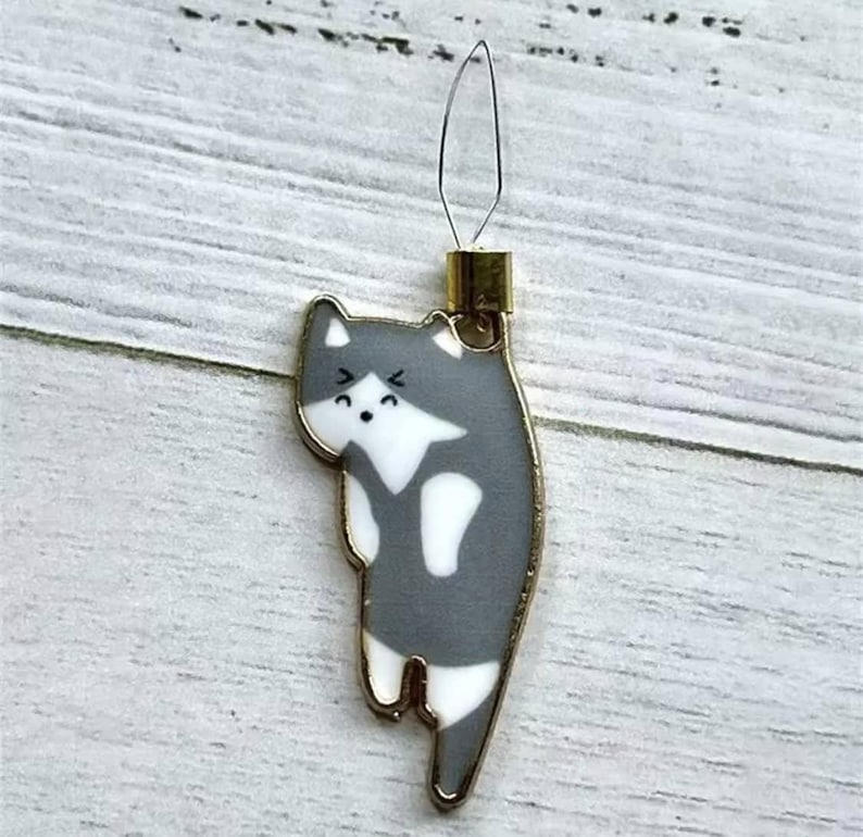May include: A gray and white cat-shaped needle threader with a gold metal loop. The cat has a white face with black eyes and a black mouth.
