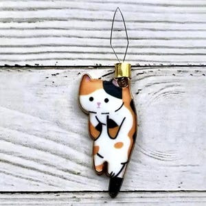 May include: A gold-colored cat-shaped needle threader with black, orange, and white markings. The cat is holding its paw up and has a cute expression on its face.