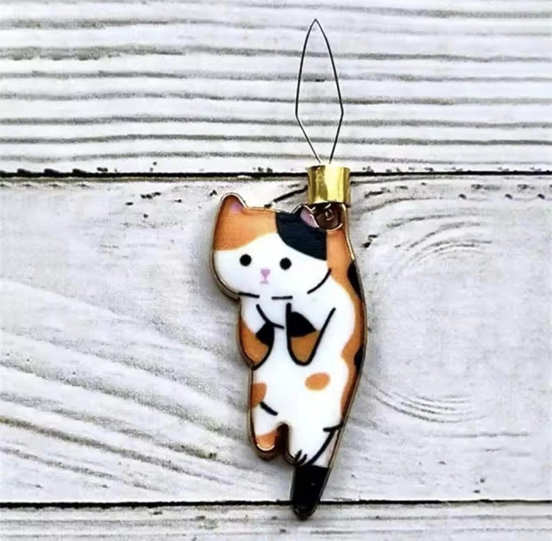 May include: A gold-colored cat-shaped needle threader with black, orange, and white markings. The cat is holding its paw up and has a cute expression on its face.