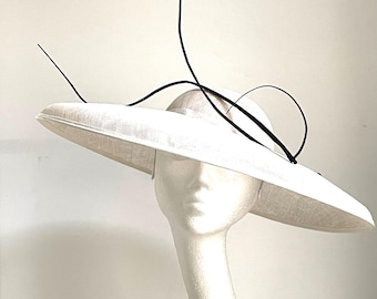 May include: An off-white hat with a wide brim and a structured crown, adorned with a black decorative element. The hat is displayed on a white mannequin head. The hat's brim is wide and flat, and the black accent adds a touch of elegance.
