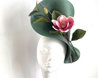 May include: A green, wide-brimmed hat with a pink flower and green leaves. The hat is on a white mannequin head.