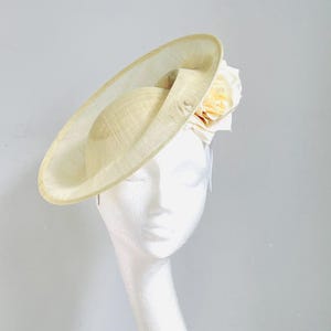 May include: A cream-coloured hat with a wide brim and a rounded crown. A large, pale yellow rose is attached to the side. The hat is displayed on a white mannequin head, showcasing its elegant design.
