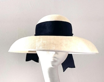 May include: A wide-brimmed cream-colored hat featuring a textured straw-like weave. The hat is accented with a wide, smooth black velvet ribbon band that encircles the crown and hangs down in two short, notched tails at the back of the brim.