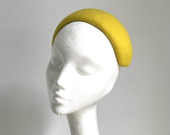 Puede incluir: Una diadema de fieltro amarillo con forma curva. La diadema está en una cabeza de maniquí blanca.