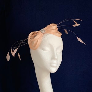 May include: A peach-coloured fascinator with a bow design and a sparkling rhinestone accent. The headpiece features delicate feather accents and is displayed on a white mannequin head against a dark blue background. This elegant accessory is suitable for special occasions.